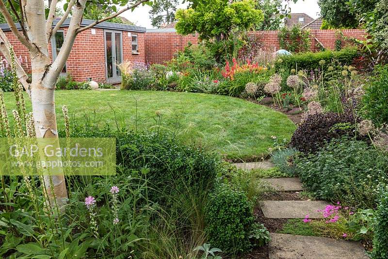 A suburban country garden with a stepping stone path leading to a circular lawn and mixed borders, small brick studio workshop in background