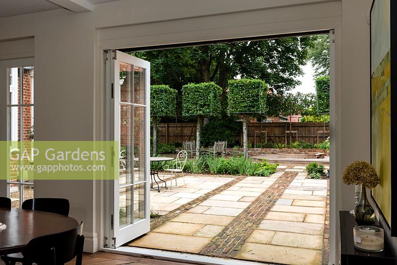 View from inside house through open doors to patio with bands of pavers and bricks to clipped Carpinus - Hornbeam - square trained trees