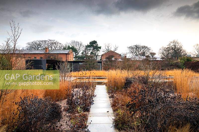 Paved path leading to pond surrounded by winter borders and grasses.