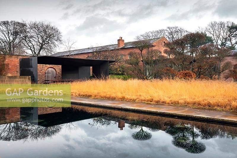 View across large pond, with reflections of trees and buildings in water, to a bed of Molinia. A modern design within an old walled garden, pavilion with seating 