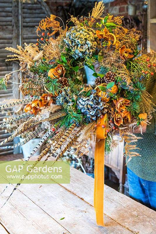 Man holding finished rustic orange, green and blue festive wreath in workshop