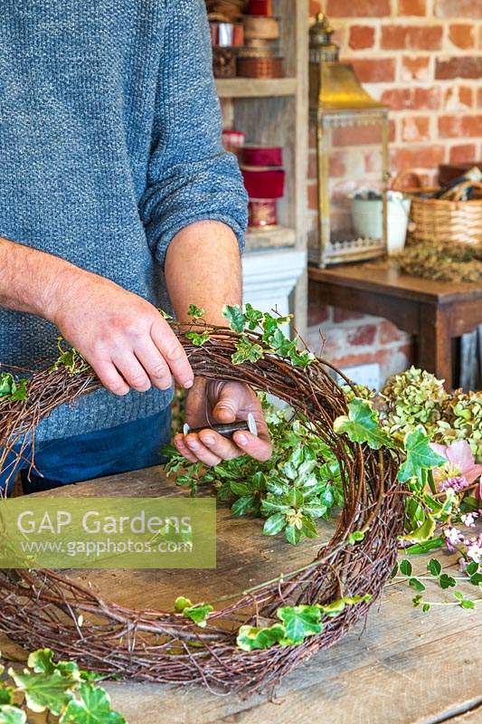 Man using floristry wire to attach ivy to rustic wreath form. 