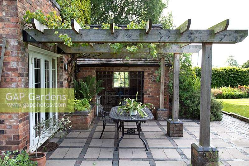Sturdy wooden pergola attached to house with Vitis vinifera - Grape Vine, underneath a patio with grid pattern of paving, with dining table and a corner raised bed in brick 