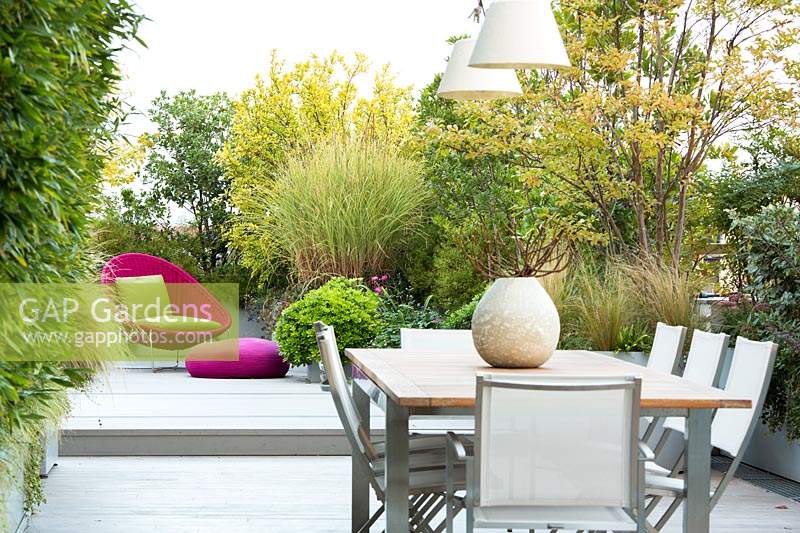 Dining area on terrace screened by container grown shrubs and relaxing area in the background with bright pink wicker chair and cushions