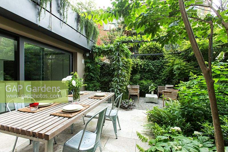 Garden outside house divided into a dining area and more relaxed seating area by metal pergola and climbers, perimeter of property screened by foliage trees and shrubs