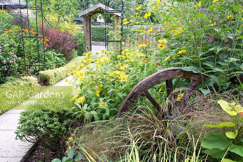 Wooden old cart wheel amongst plants in a border, view of path and obelisks beyond