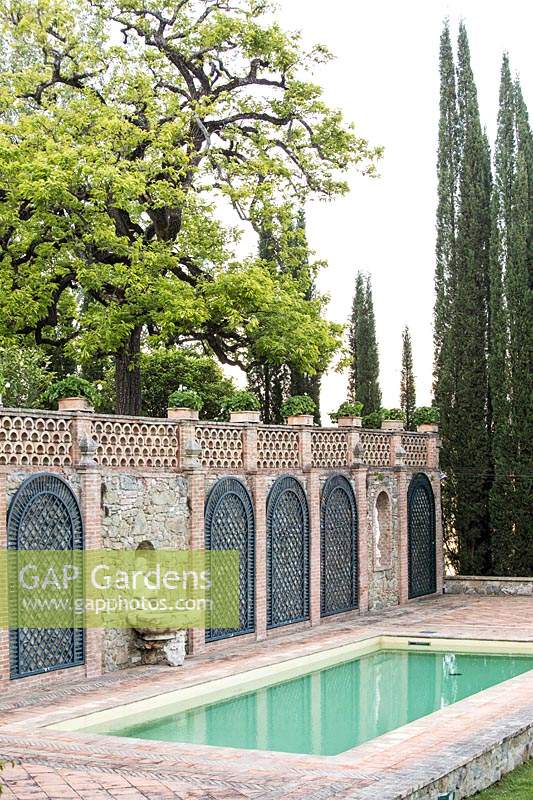 Swimming pool set in terrace with wall with trellis panels and alcove statue, Cupressus sempervirens - Cypress - trees