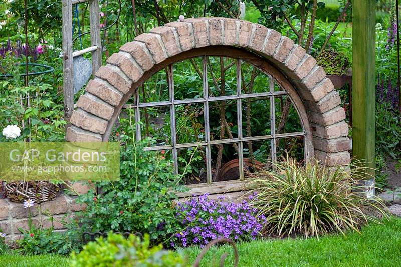 A window from a flea market framed by brick, used as divider or screen in the garden with low plants in front