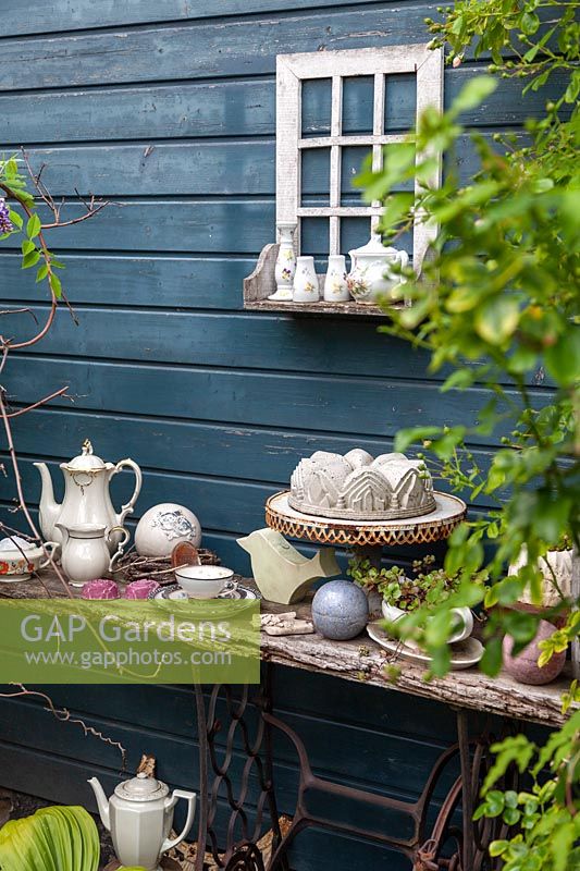 A collection of eclectic objects: tea cups, coffee pots, a cake plate and sauce boat, displayed on old timber shelf near veranda wall