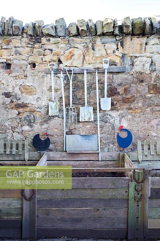 Garden tools hanging on stone wall above a row of compost bins 