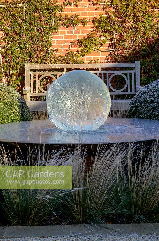 Aqualens water feature designed by Allison Armour with large clipped balls of Buxus sempervirens, Stipa tenuissima grass and Traschelospermum jasminoides on brick wall, on a frosty morning.