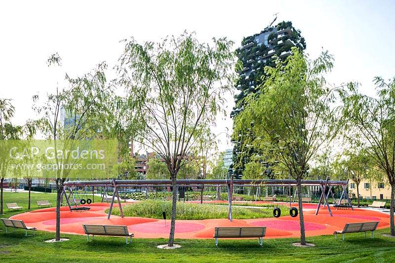 View across public park with young trees and benches around a playground, in background the Bosco Verticale - Vertical Forest - by architect Stefano Boeri 
