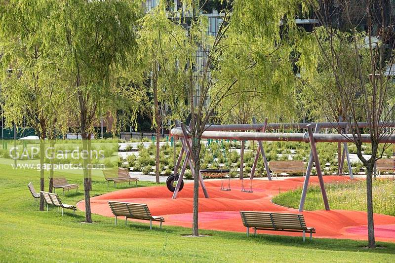 View of public park with young trees, benches and playground surrounded by planting and grass