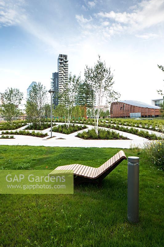 Public park with seating on grass and recent plantings of young trees, geometric beds with paths 