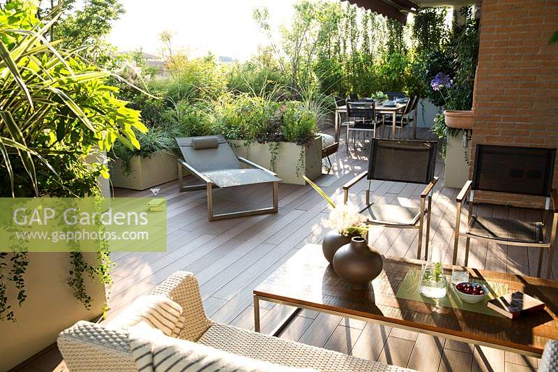 The living area of the terrace with relaxed seating and sun lounger in front of planter with mixed foliage, in foreground a planter with Phormium and other plants provide a screen 