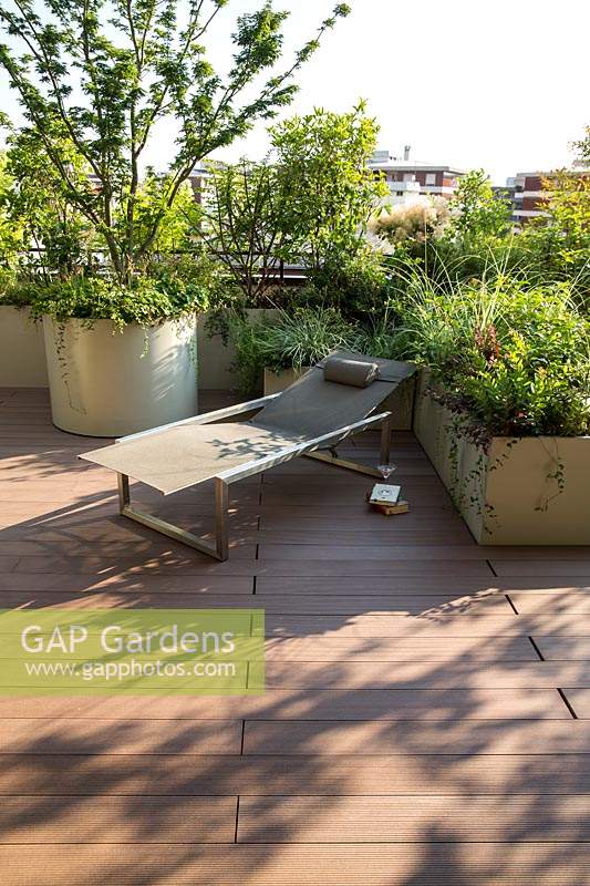 View over decked terrace to sun lounger with containers providing a screen from edge. Round container with Acer palmatum 'Crispifolium', other trough planters with foliage perennials.