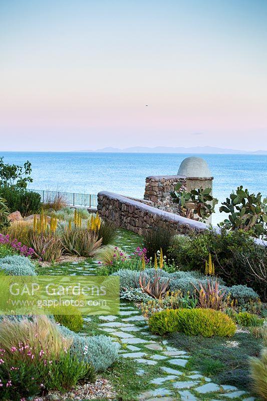View over terrace wall to the sea. Planted with drought tolerant and wind resistant planting, mixing Mediterranean shrubs: Pistachia lentiscus, Myrtus communis, Helichrysum with plants from similar climates:  This Aloe and Agave, Stipa tenuissima, Muhlenbergia capillaris, Chondropetalum tectorum and Tamnochortus insignis