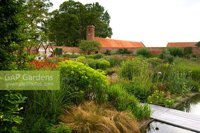 Modern formal country garden, view over deep flower beds with decked path to walls and buildings 