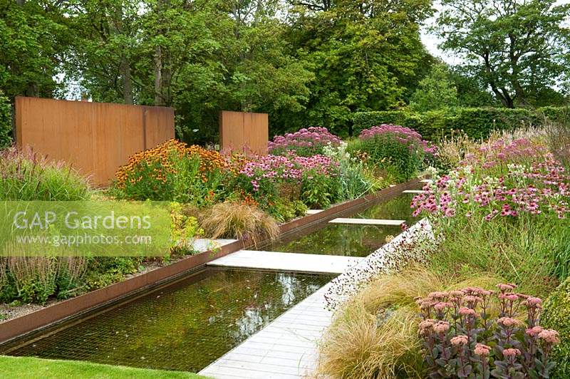 Modern water feature with low bridge paths, surrounded by perennial flower beds with a background of corten steel and trees