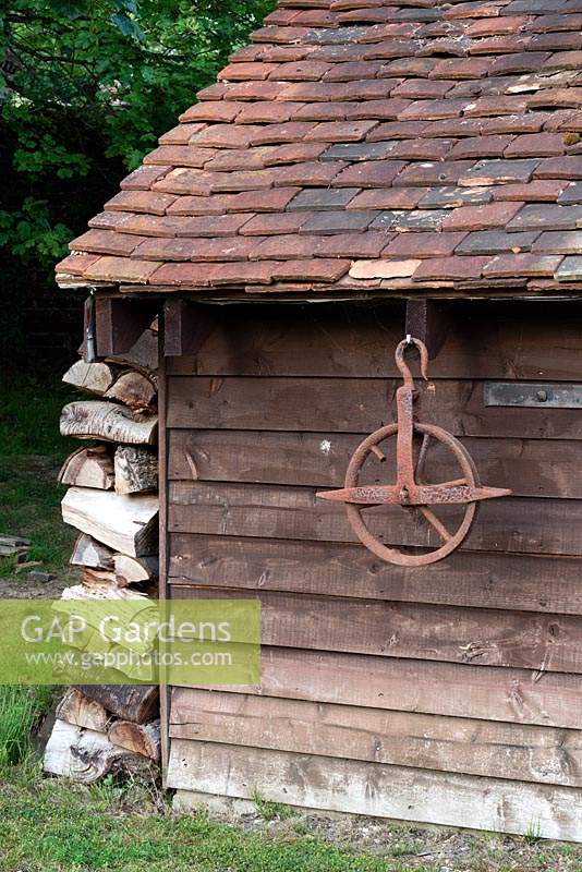 Old wooden shed with tiles roof, log pile and an old pulley wheel hanging from the roof
