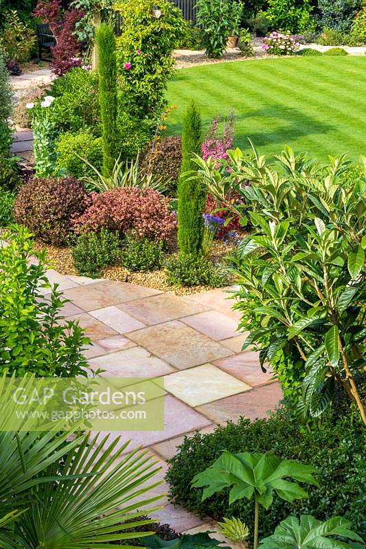View across a garden with wide paved pathways which encircle a small striped lawn. Beds planted with neat shrubs such as pencil conifers and Hebe
