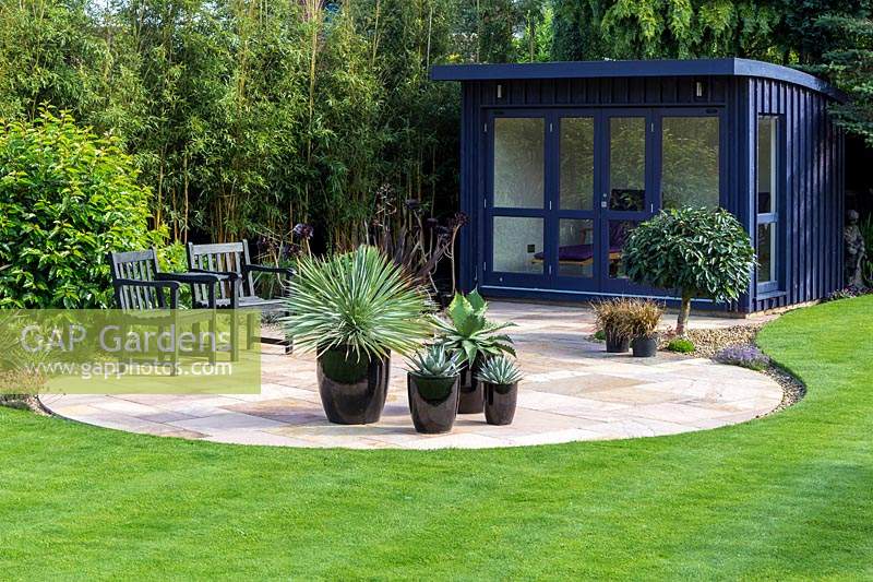 View over neat lawn and patio to a hand-built garden room. Agaves and a young Cordyline in black ceramic pots on the circular patio, Bamboo screen behind summerhouse.
