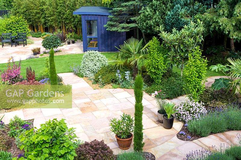 View over paved area to the blue painted garden room with curved roof. Plants in beds: Trachycarpus fortunei - Palm, Eriobotrya japonica, Tetrapanax papyrifer, Lavandula - Lavender, variegated Pittosporum, pencil conifers and Hebes 