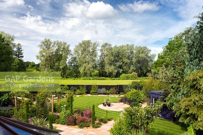 Overview of the garden with circular patios, lawn, beds of shrubs and black painted fence at boundary