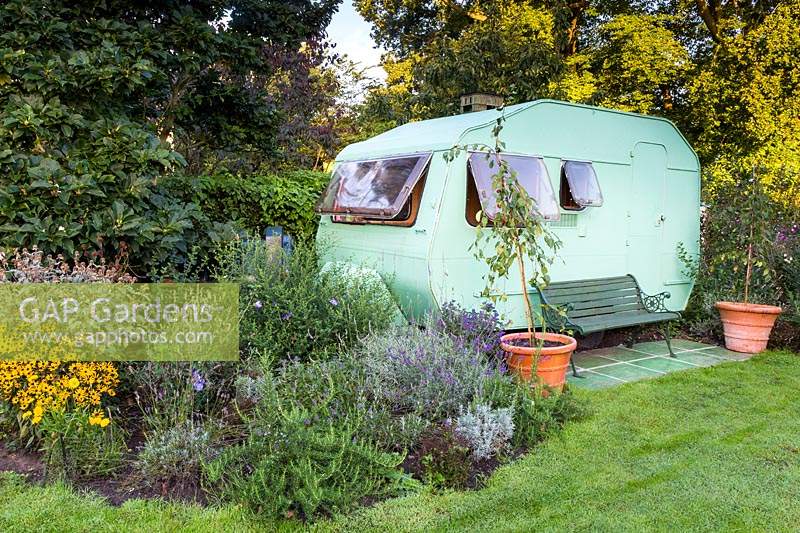 Super Sprite caravan which serves as a home office, nearby paved area for seating and young Malus 'Red Jade' - Crabapple - in terracotta pots. Also a mixed bed of perennials include Rudbeckia fulgida var. sullivantii 'Goldsturm', Salvia rosmarinus - Rosemary, Lavandula x intermedia 'Grosso'  - Lavender and Caryopteris clandonensis