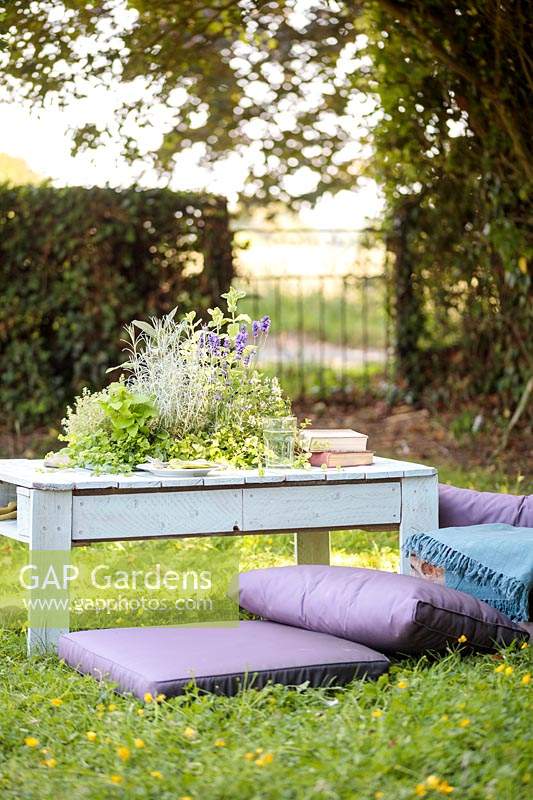 Table made out of a wooden pallet with central planter planted with mixed herbs in summer country garden with cushions