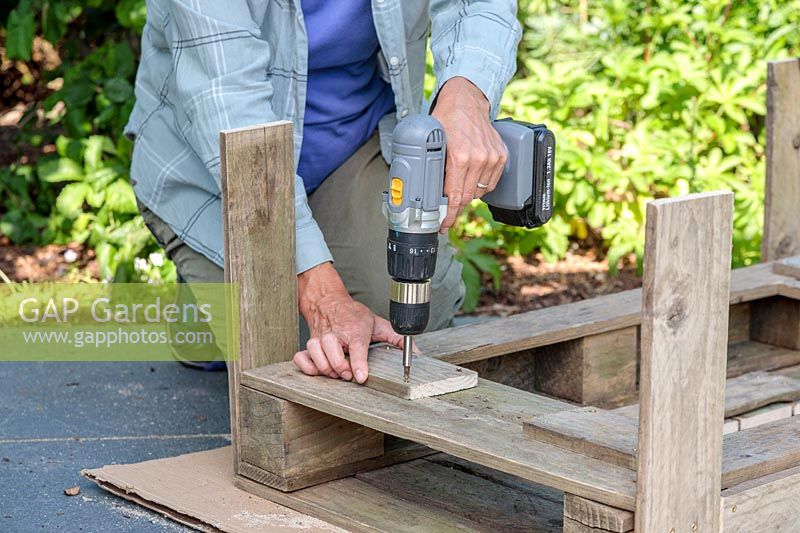 Woman using a cordless electric screwdriver to attach  wood to make a shelf under the table. 