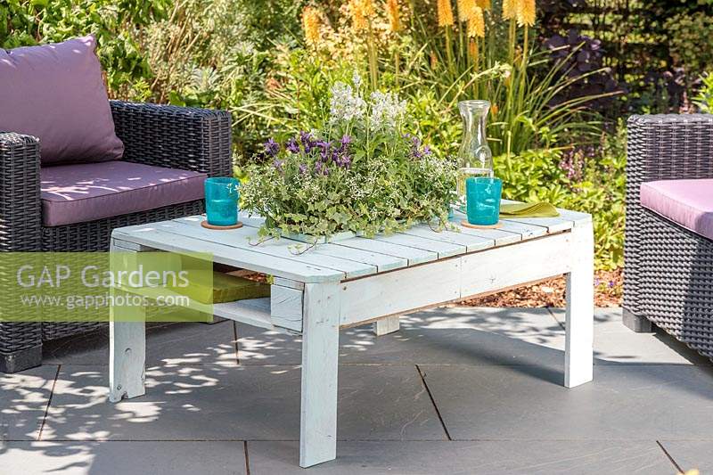 Wooden pallet table with intergral sunken planter, with summer planting on slate patio in summer, accompanied by outdoor chairs.