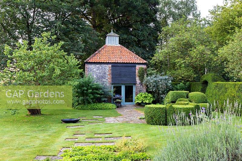 View of the 18th century dovecote in a garden setting: Buxus sempervirens - Box - topiary, a mosaic of paving stones in lawn Cercis siliquastrum - Judas Tree. Near dovecote a pot of Hosta 'Sum and Substance' and Rosa glauca. 
