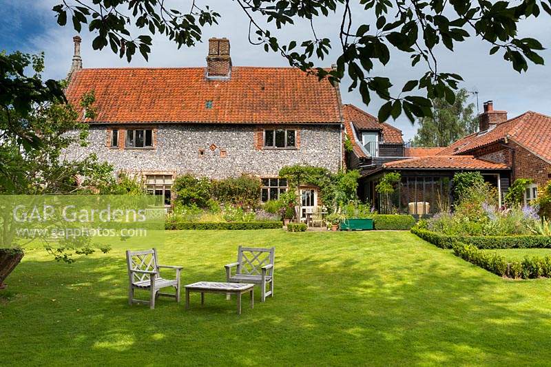 View across large lawn with wooden seats, towards house and terrace edged with Buxus - Box