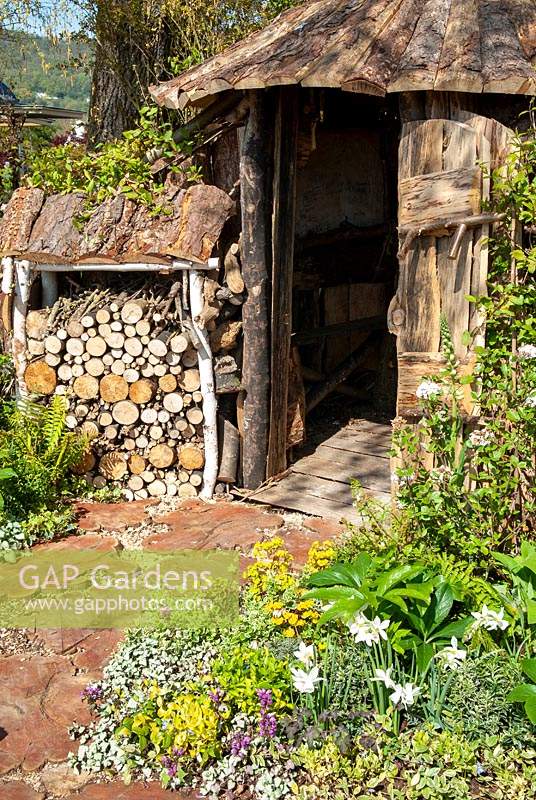 Path of tree trunk slices and wood chips, leading to rustic wooden hut and wood store - RHS Malvern Spring Festival
