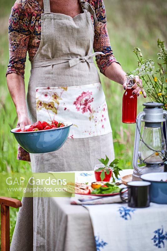 Woman wearing floral apron adding fresh fruit and drink to picnic table