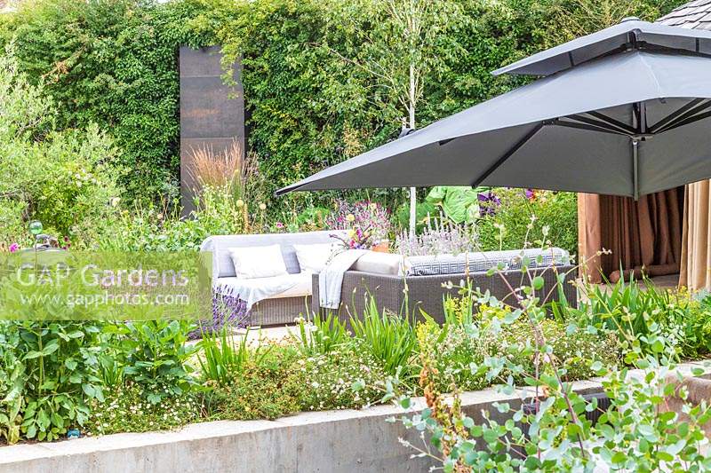 View under parasol to patio with lounge furniture surrounded by flower beds