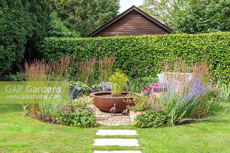 Stepping stone in lawn leading to paved area with corten steel water feature and circular planting