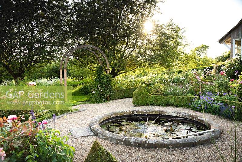 Circular pond with water spouts surrounded by box-edged beds of roses 