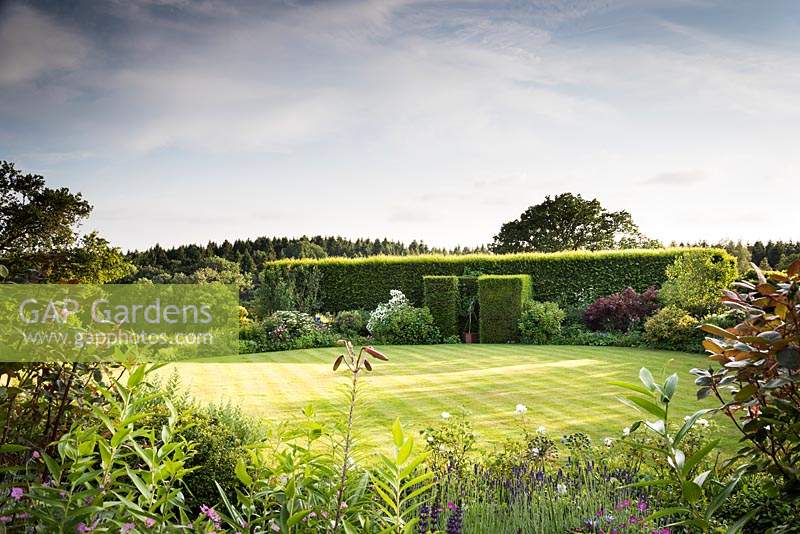 Circular lawn surrounded by borders of shrubs and herbaceous perennials, blocks of yew framing a figurative sculpture and a line of hornbeam 