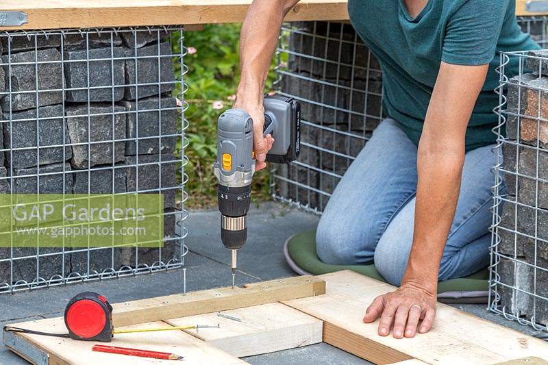Woman using an electric screwdriver to screw through a batten, to construct a table top made up of cut scaffolding boards. 