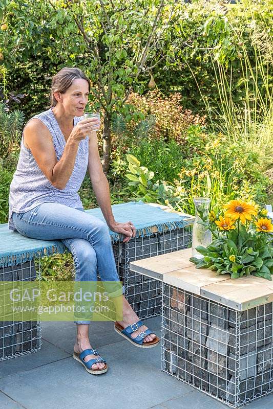 Woman sitting and enjoying a drink on gabion and scaffolding board bench that is complimented by a matching table on slate patio in modern garden. 