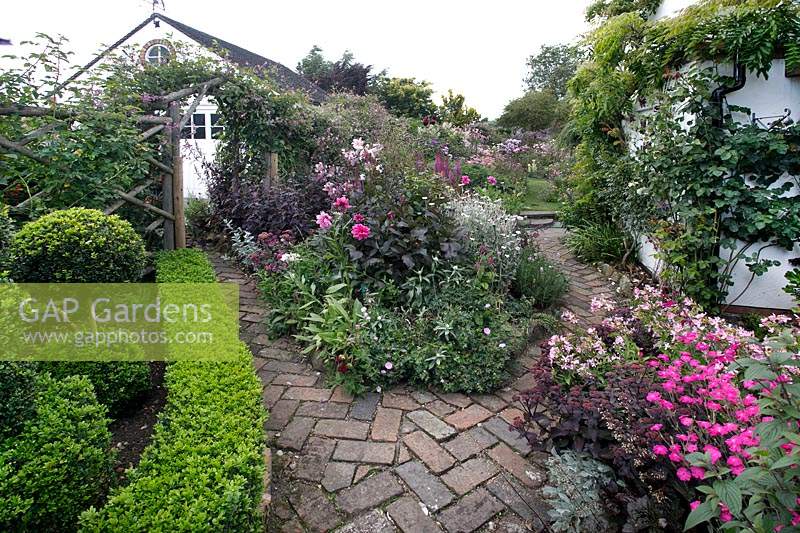 Front garden with brick paved paths, to one side topiary, box hedging and rustic arch lead to garage, to the other full flower beds and side of house with climbers