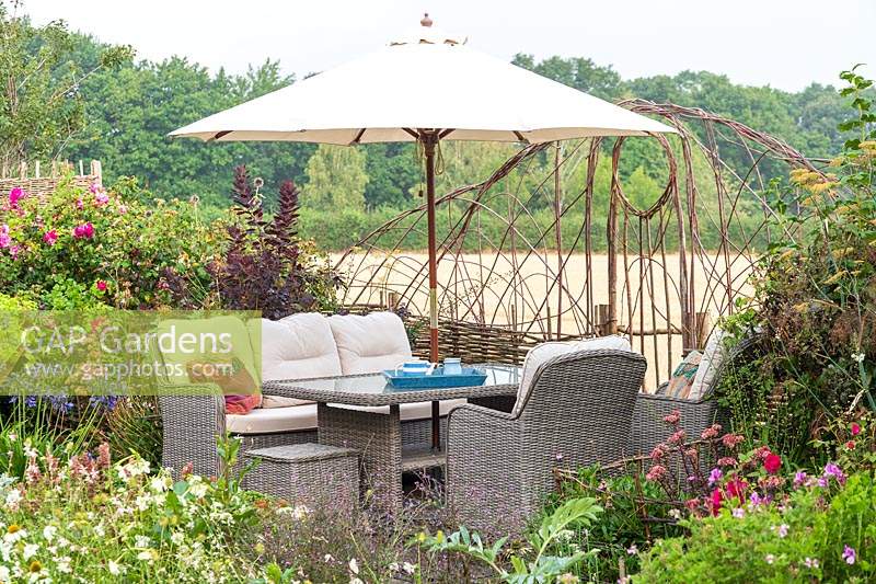 Seating area with lounge garden furniture and parasol, view through woven hurdle to countryside 