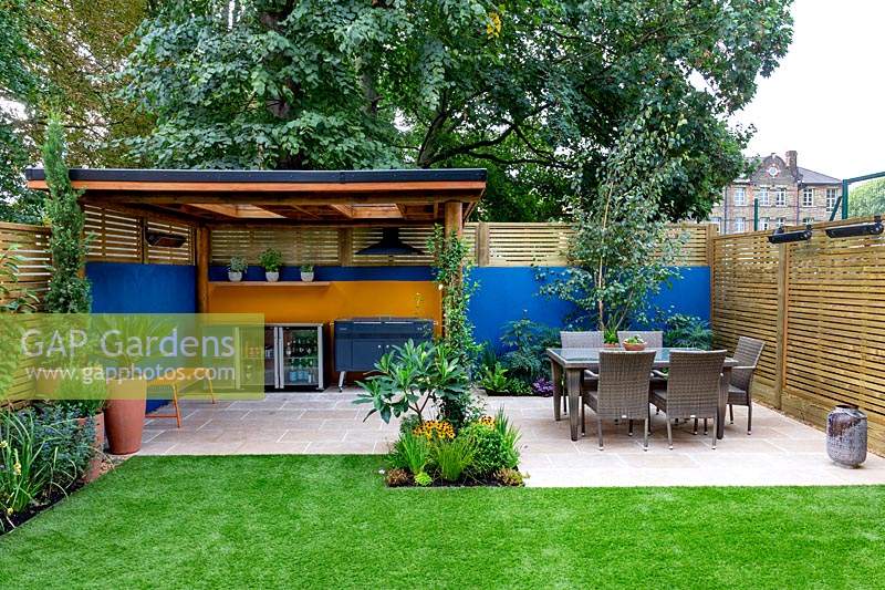 Patio, table and chairs and outdoor room with covered barbecue. Surrounded by a contemporary wooden trellis fence and blue painted wall.