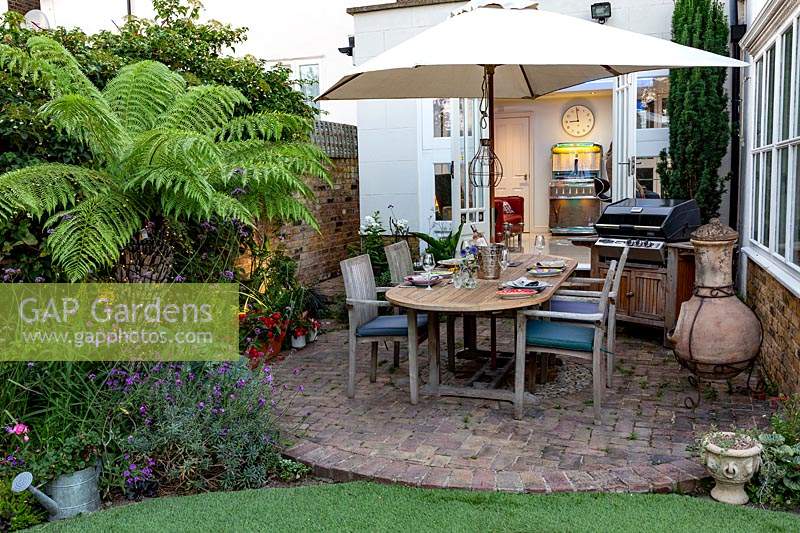 Small garden patio with table and parasol, barbecues and Dicksonia antarctica - Tree Fern - in bed near lights