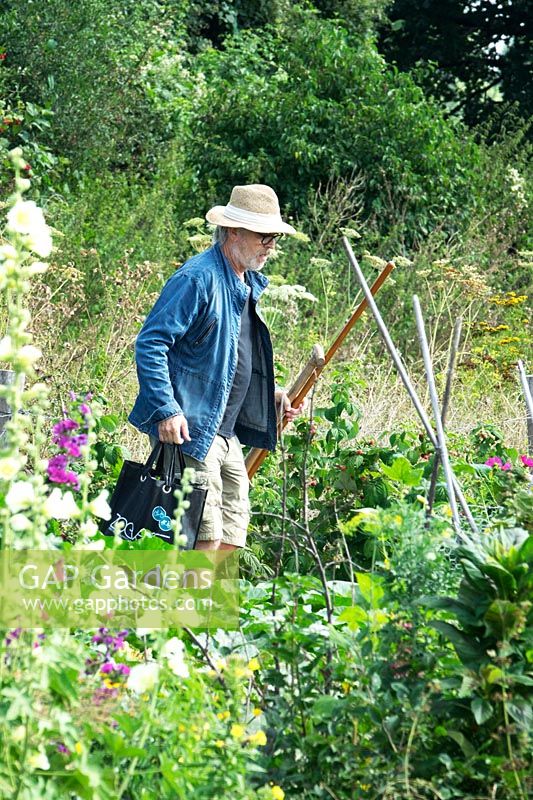 Man wearing sun hat, walking to his plot carrying tools and a bag for harvesting 