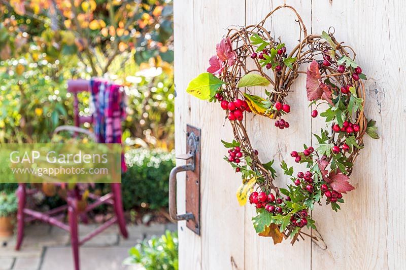 Woven heart-shaped wreath with berries and foliage, hanging on open wooden door, garden beyond 