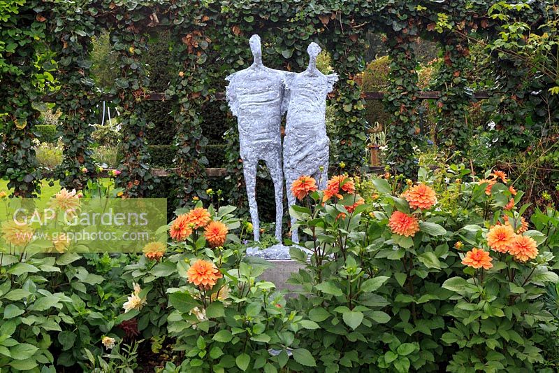 Sculpture in the manner of Giacometti of two people together, amongst Dahlia 'Nicolas'. A trellis with round-leaded ivy, Hedera sp. behind.