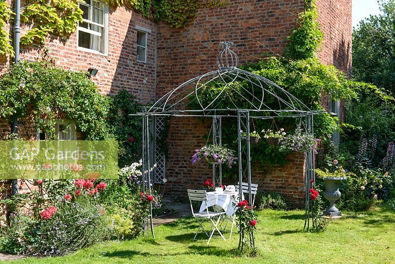 Gazebo on lawn by brick house, gazebo has Rosa - Rose - climbers trained up it and hanging baskets with seating underneath
